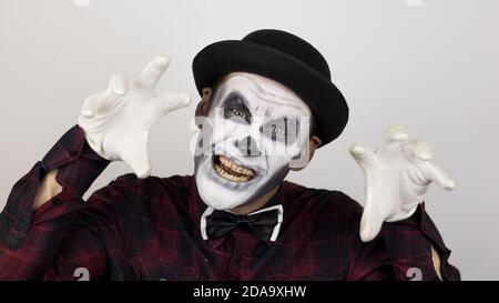 A horrible man in clown makeup grimaces and makes frightening gestures. A scary clown looks at the camera and laughs terribly. Stock Photo