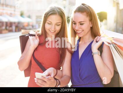 Young happy millennial friends walking on streets with shopping bag and checking social media platform updates - Two students ordering clothes online Stock Photo