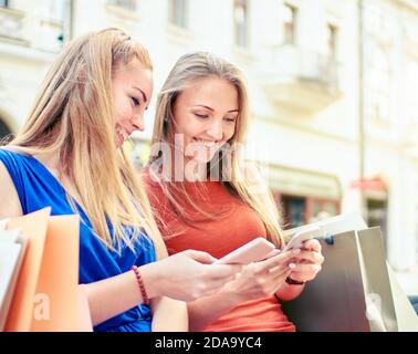 Young smiling millennial friends sitting on bench with shopping bags around and checking social media platform updates - Two students ordering clothes Stock Photo