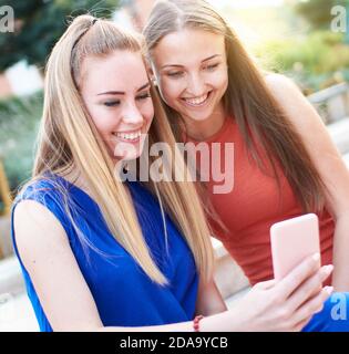 Smiling, young millennial female friends checking social media updates on smartphone - Two happy woman sitting on bench and ordering shoes from websho Stock Photo