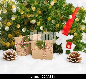 Two handmade plastic-free Christmas presents, wooden reindeer in Santa hat and cones in snow with pine tree branches and bokeh lights as background. W Stock Photo