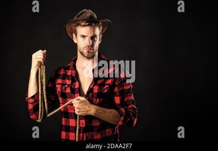 Lasso tool of American cowboy. Still used on ranches to capture cattle or other livestock. Western life. Man unshaven cowboy black background. Cowboy wearing hat hold rope. Lassoing on prairie. Stock Photo