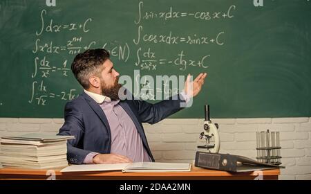 Teacher charismatic hipster sit at table classroom chalkboard background. Educational conversation. Talking to students or pupils. School teacher concept. Teacher bearded man tell interesting story. Stock Photo