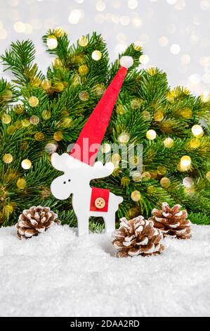 White Christmas reindeer in red Santa hat and pine cones in snow with fir tree and glowing bokeh lights as background. Festive holiday greeting card. Stock Photo