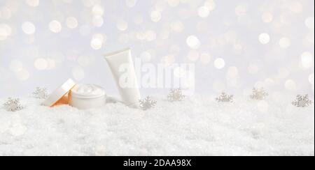 Winter skin care cosmetic products in snow and snowflakes on white background with bokeh lights. Opened face cream jar and hand creme or body lotion Stock Photo