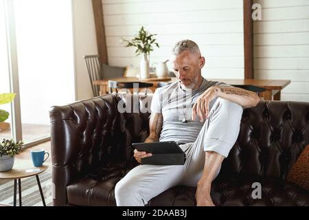 Focused middle aged caucasian man in casual clothes using digital tablet while sitting on sofa in the living room of his modern apartment Stock Photo