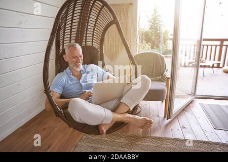 Cheerful middle aged caucasian businessman in casual clothes sitting with laptop on swing chair at his modern apartment, working from home Stock Photo