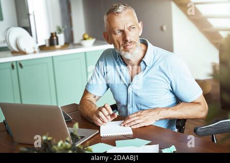 Home office. Focused middle aged caucasian man making some notes while sitting at table and working online Stock Photo