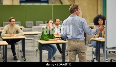 Business education. Rear view of a male business coach or speaker pointing at flip chart and giving presentation to audience, Stock Photo