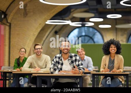 Young cheerful mixed race male office worker sitting with collegues in coworking space and listening to coach or speaker during business seminar, selective focus on man. Education and self improvement at work Stock Photo