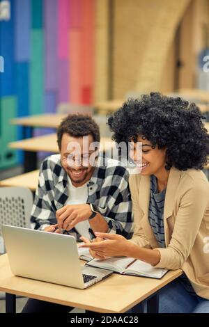 Two young happy diverse colleagues looking at laptop and discussing something while sitting at the desk in coworking space. Teamwork and collaboration concept Stock Photo