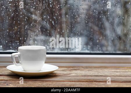 White steaming cup of hot tea or coffee on vintage wooden windowsill or table against window with raindrops on blurred background. Shallow focus. Stock Photo