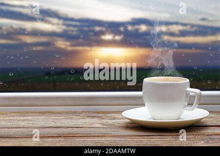 White steaming cup of hot coffee on vintage wooden windowsill or table against window with raindrops and sunset on blurred background. Shallow focus. Stock Photo