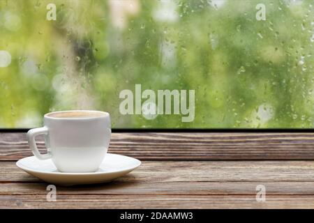 White steaming cup of hot coffee on vintage wooden windowsill or table against window with raindrops and foliage on blurred background. Shallow focus. Stock Photo