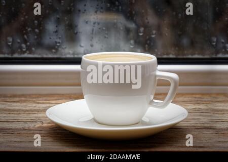 White ceramic cup of hot coffee on vintage wooden windowsill or table against window with raindrops on blurred background. Shallow focus. Stock Photo