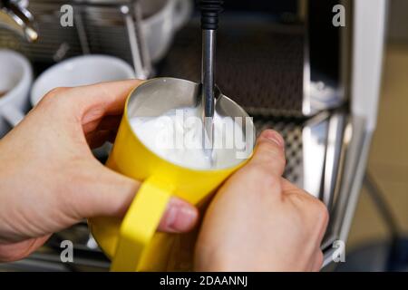 Closeup hands of barista who steaming milk for cappuccino or latte in yellow pitcher. Shallow focus. Stock Photo