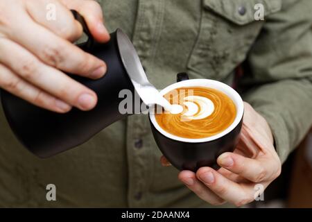 https://l450v.alamy.com/450v/2daanx4/barista-holding-a-black-cup-in-one-hand-and-a-black-pitcher-in-other-and-pouring-milk-into-a-cup-of-cappuccino-to-make-latte-art-shallow-focus-2daanx4.jpg