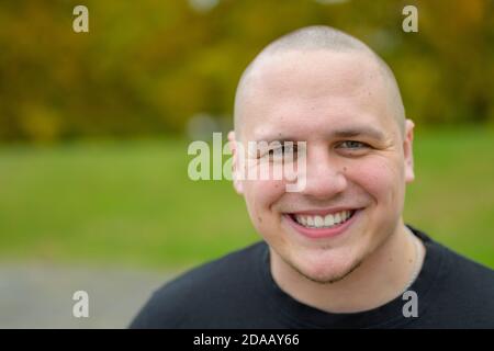 Young man with shaved head standing outdoors in a park with a pleased smile and thoughtful expression Stock Photo