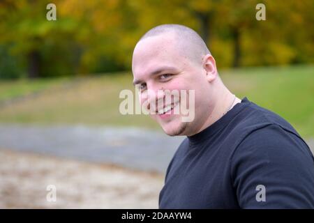 Smiling friendly thickset young man with a shaved head looking sideways at the camera outdoors in a park in autumn Stock Photo