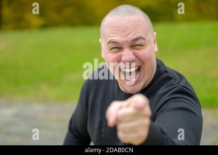 Emotional young man laughing or shouting at the camera while pointing his finger with focus to his face outdoors in a park Stock Photo