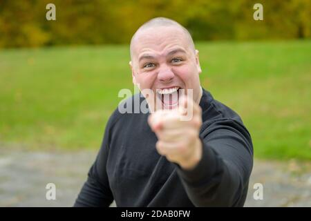 Emotional young man laughing or shouting at the camera while pointing his finger with focus to his face outdoors in a park Stock Photo
