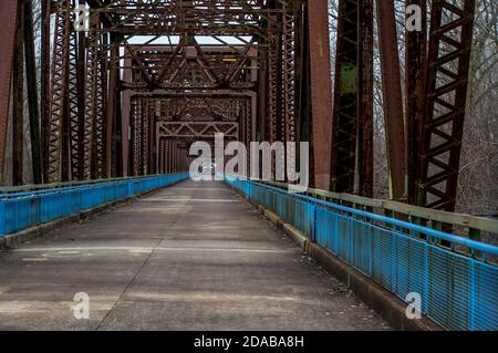 Old rusty steel truss bridge going into background with concrete deck and blue rails for pedestrians in winter Stock Photo