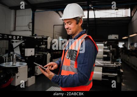 Smiling male engineer wearing uniform scrolling on tablet standing in factory Stock Photo