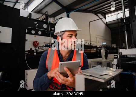 Male engineer wearing hard hat researching technical drawings on tablet while sitting in factory. Stock Photo