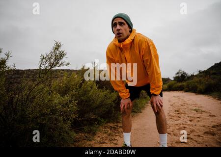 Close up tired young male athlete resting to catch breath after downhill sprinting on gravel trail in cloudy weather with foliage Stock Photo