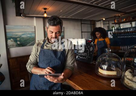 Male waiter scrolling on digital tablet searching for menu while waitress makes coffee in coffee restaurant Stock Photo