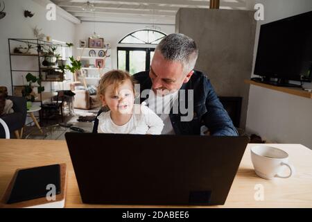 Father and daughter watching family videos on laptop drinking coffee. Stock Photo