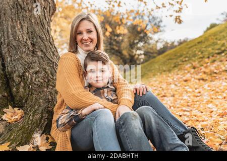 Happy family mother and child boy in the autumn leaf fall in park sit on the bottom of the tree Stock Photo
