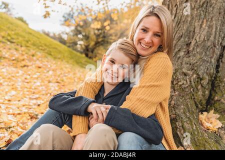 Happy family mother and child boy in the autumn leaf fall in park sit on the bottom of the tree Stock Photo