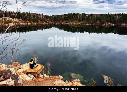 young man from the back in warm clothes relaxing on the rocky shore of the lake. Beautiful autumn lake landscape. Hiking alone Stock Photo