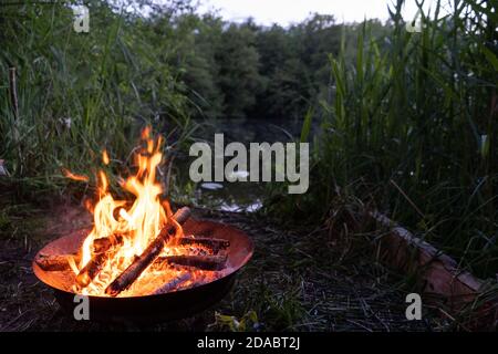 Fire pit with flames and burning logs next to romantic lake and fire in fire pit at dusk in campsite, boho atmosphere Stock Photo