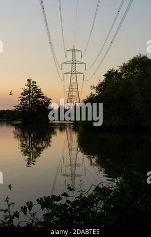 Silhouetted pylon and one flying bird reflected onto beautiful still peaceful lake, at dusk in summertime England. An orange hint of colour, last glim Stock Photo