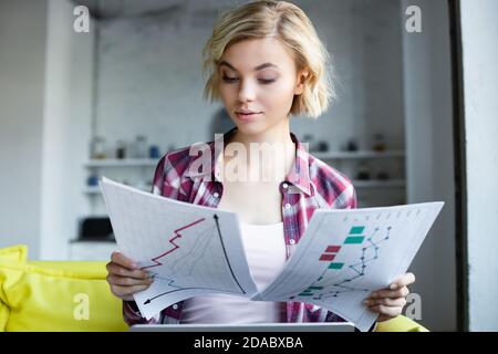 blonde woman in checkered shirt working from home and looking at graphs Stock Photo