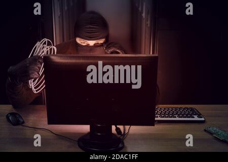 A hacker in a black mask with wires in his hand in a server room. Concept of data security problems and network hacking Stock Photo