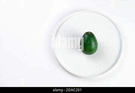 Green unpeeled avocado in a flat white plate on a light background. Stock Photo