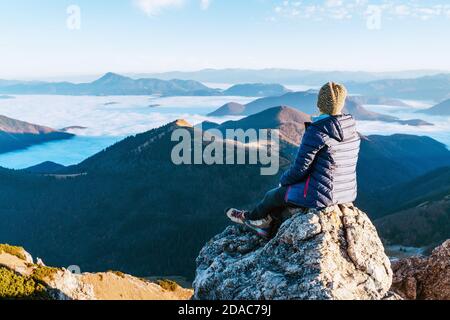 Young hiker woman sitting on the mountain summit cliff and enjoying mountains valley covered with clouds view. Successful summit climbing concept imag Stock Photo