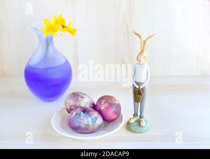 Cozy Easter holiday composition with cute bunny, chicken eggs painted in galaxy colors and vase with narcissus flowers. Stock Photo