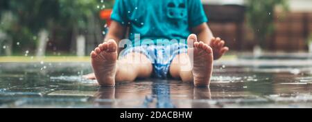 No face photo of a caucasian small boy in blue clothes and barefoot sitting on the ground in water and playing