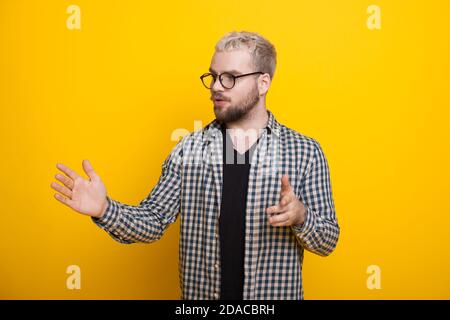 Caucasian man with beard and blonde hair is explaining something on a yellow studio wall gesturing with hands and wearing glasses Stock Photo