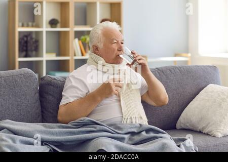 Sick senior man with bad cold or flu sitting on sofa at home and taking medicine with water Stock Photo