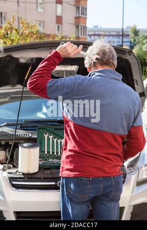 Rear view at confused mature man scratching the back of his head with wrench while standing behind opened hood of car, looking at breakdown engine Stock Photo