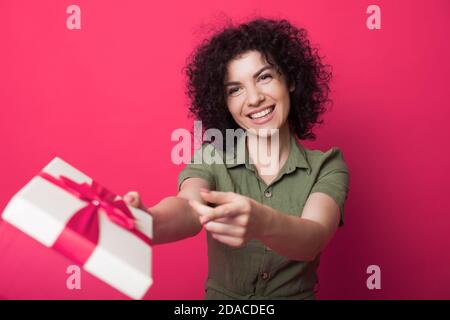 Adorable woman with curly hair is is receiving a present on a red studio wall smiling and holding a packed gift box Stock Photo