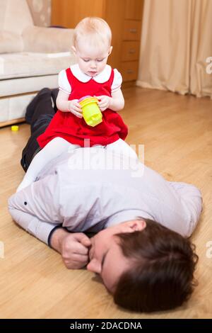 Little daughter sitting on the back of tired father lying unconscious on floor, man sleeping after playing toys with his child Stock Photo