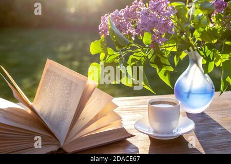 Little white cup of espresso coffee, opened book, blue semi-transparent vase with purple lilac flowers on rustic wooden table in the garden Stock Photo