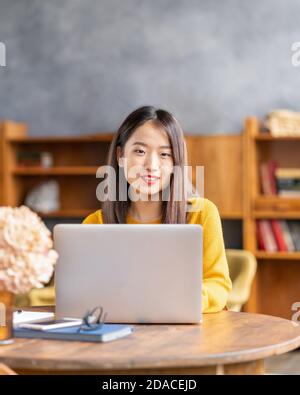Happy smiling asian woman working on laptop at home or in cafe. Young lady in bright yellow jumper Stock Photo