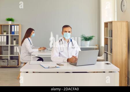 Male doctor in protective medical mask sitting at workplace with computer in office. Stock Photo
